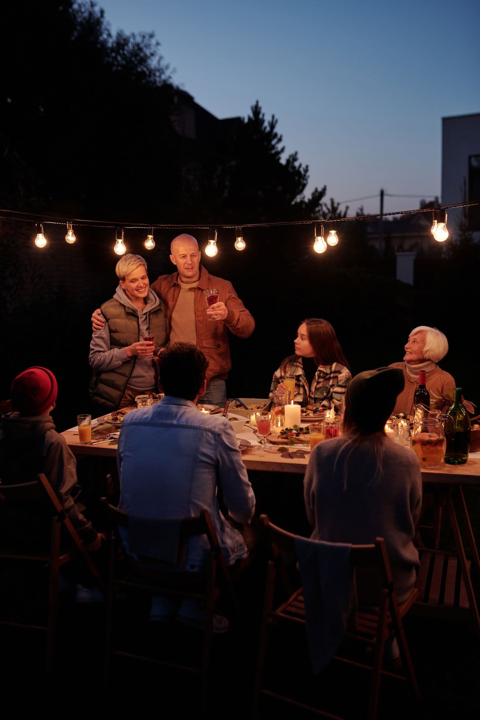 A multi-generational family sharing dinner together outdoors at dusk in a warmly lit backyard